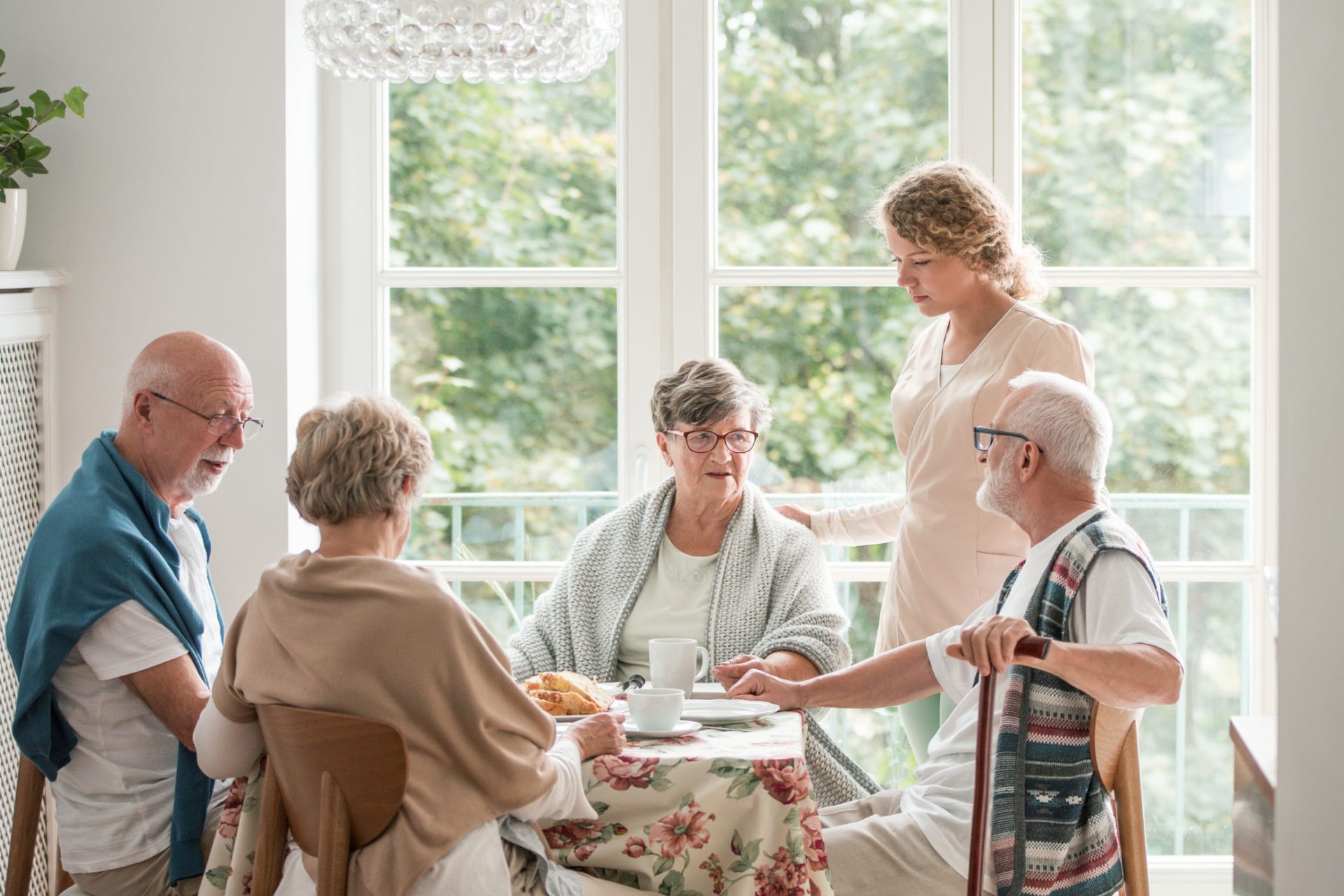 Group of senior friends with helpful carer sitting together at the table at nursing home dining room